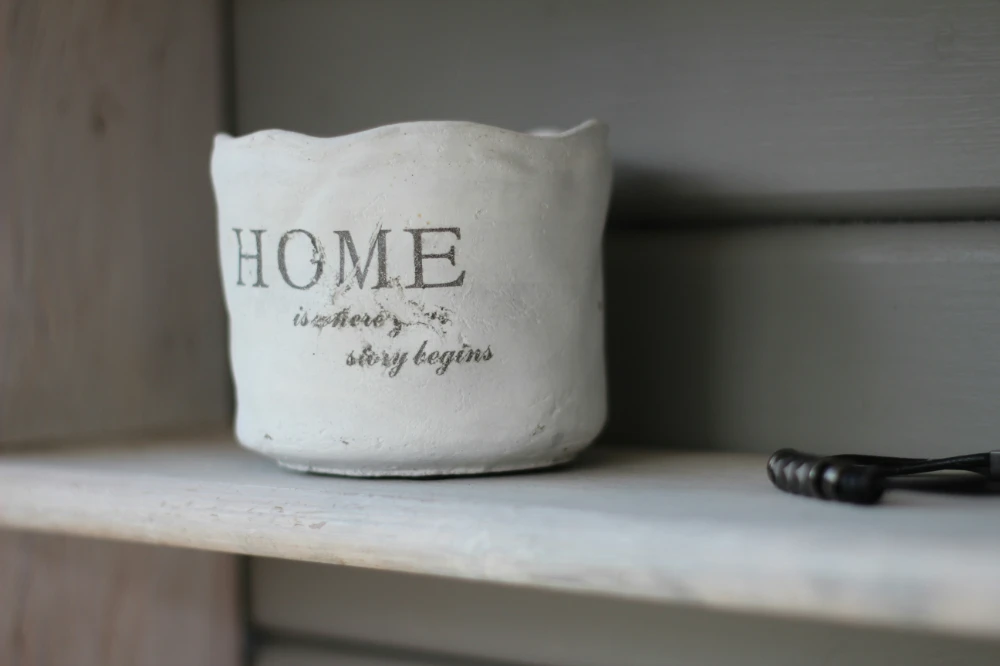 Immobilien-LUV-LEE Ein Regal mit einem hellen Tontopf auf dem HOME is where your story begins steht. Perfekt für Ihre Immobilien
